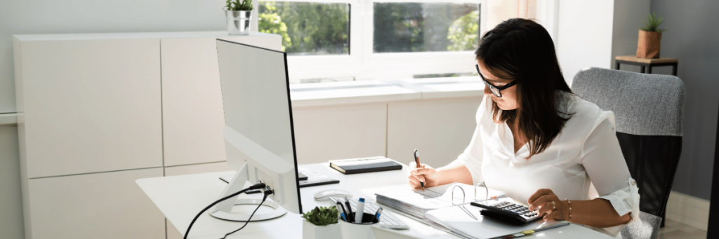 Mulher branca anotando algo em um caderno em frente a um computador.