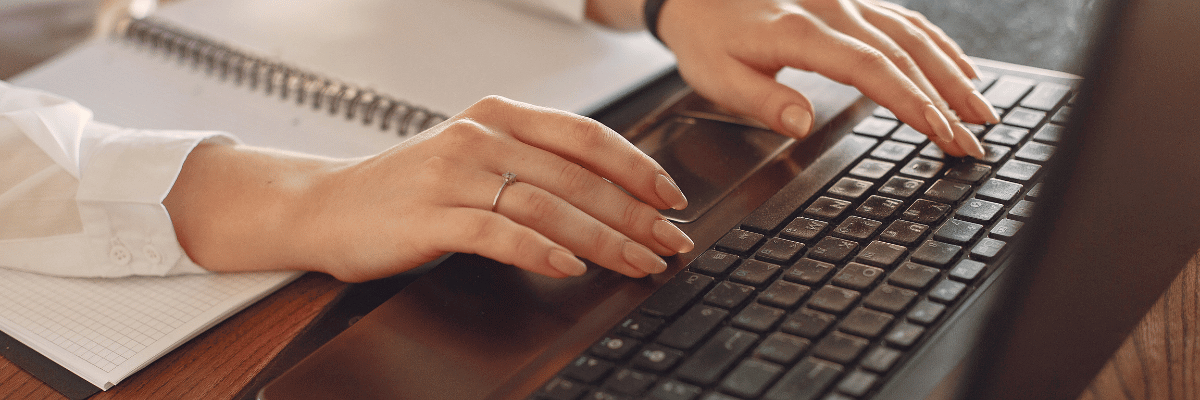 Mãos femininas digitando em um teclado preto de computador e um caderno aberto entre os dois braços