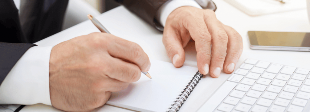 imagem mostra mãos de homem branco de terno, com uma caneta, escrevendo em uma agenda e teclado de computado a sua frente