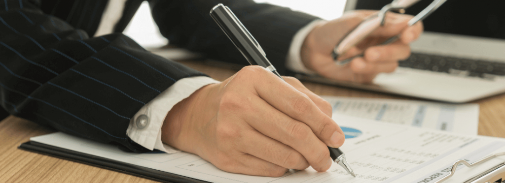 homem de terno preto com listras azuis, escrevendo em documentos em cima de uma mesa de escritório, com um óculos na outra mão e no fundo um notebook aberto.